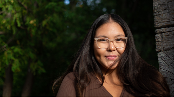 An Indigenous woman faces the camera while leaning on a wooden wall with greenery in the background