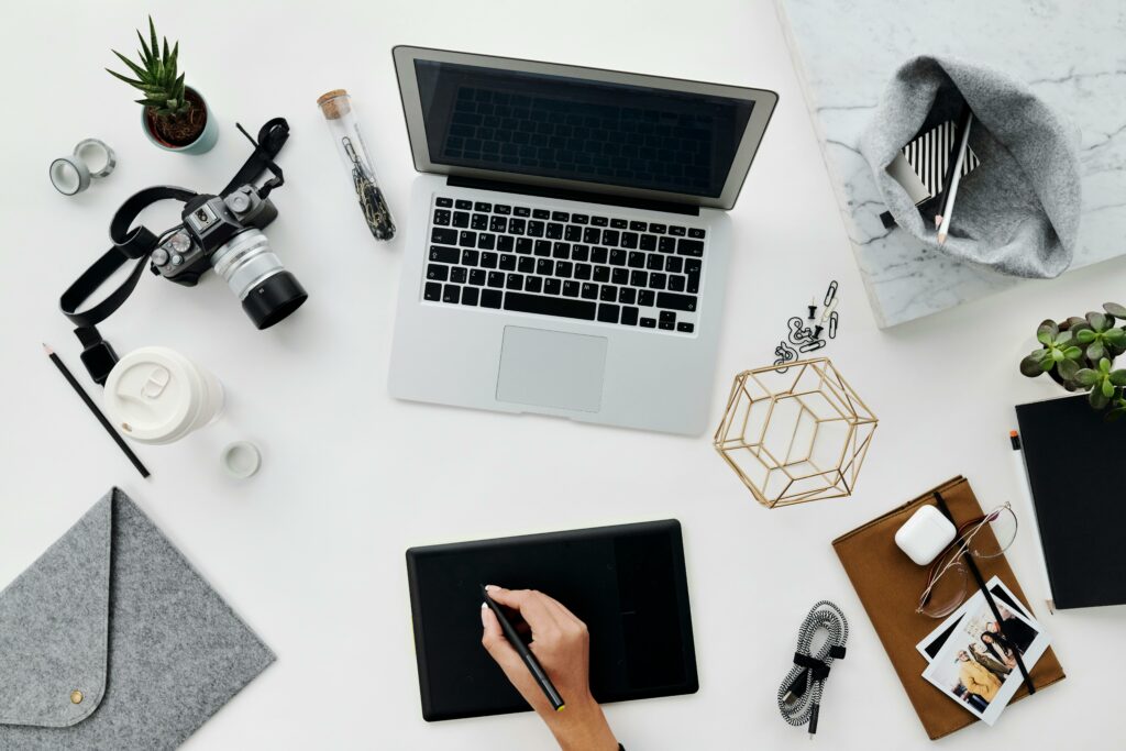 Une personne utilise un stylet et une tablette sur un espace de travail comprenant un ordinateur portable, un appareil photo, une tasse à café, des lunettes, un décor géométrique, un ordinateur portable, un smartphone et une plante succulente sur un bureau blanc.