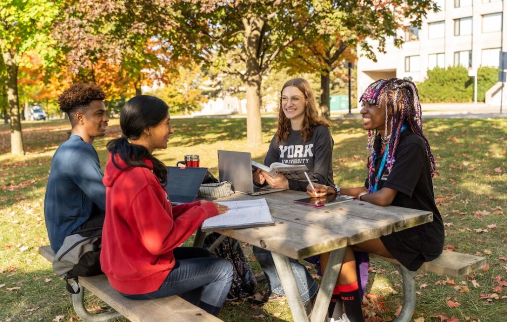 a photo of students sit at a table outside in nice weather on a York University campus