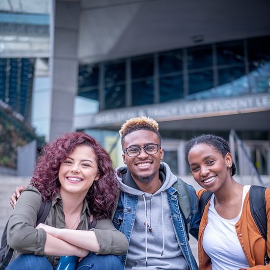 A small group of three University students sit outside on campus as they pose for a portrait. They are dressed casually in fall attire and smiling as they huddle in closely.