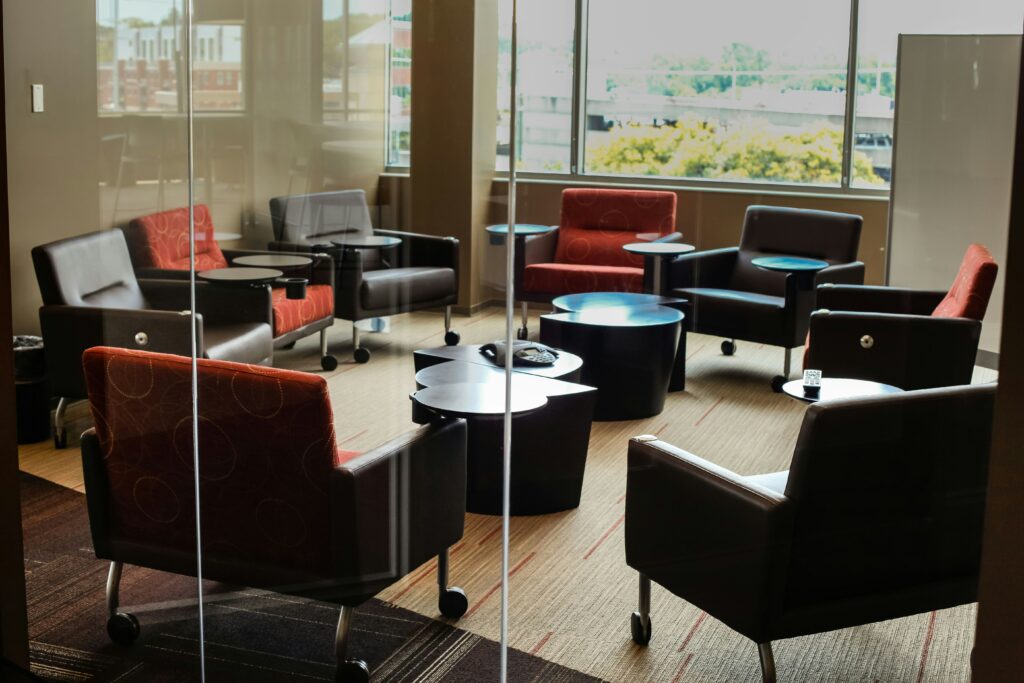 A photo of a group of fabric and leather chairs facing each other in a circle. The chairs are in a glass carpeted room, with a window looking out to trees and a parking garage. There are a few semi-circle tables in the middle of the circle. The photo is by John Price on Unsplash.