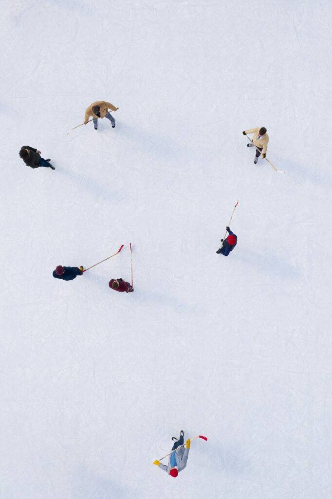 A photo of seven people playing a game of ice hockey. The background is the sheet of white ice and light snow that they are playing on. The photo is taken from a top down angle, and the camera is high above the players. The photo is by Martin Reisch on Unsplash.