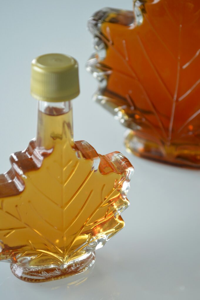 A photo of two glass maple leaf bottles filled with maple syrup. The cap is gold. There is a smaller bottle in front of a larger bottle in the back. The front bottle is focused in the shot, while the bigger bottle is unfocused in the back. They are placed on a white table which is the background of the photo. The photo is by Nadine Primeau on Unsplash.