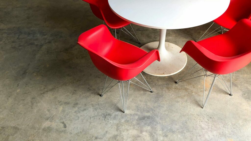 A photo of a white round table with four red chairs around it. The table and chairs are in the the top right corner of the image, with half of the table and the halves of two red chairs being cropped out of the photo. The floor is concrete. The photo is by No Revisions on Unsplash.