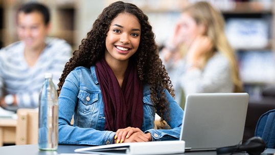 A female student sits at a desk and smiles at the camera