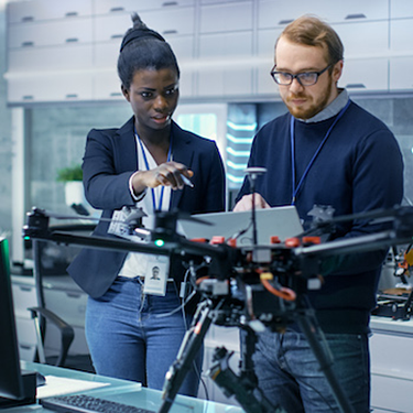 Caucasian Male and Black Female Engineers Working on a Drone Project with Help of Laptop and Taking Notes. He Works in a Bright Modern High-Tech Laboratory.