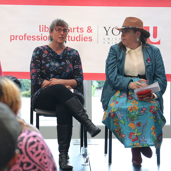 Two people sitting on chairs on a stage, in conversation.