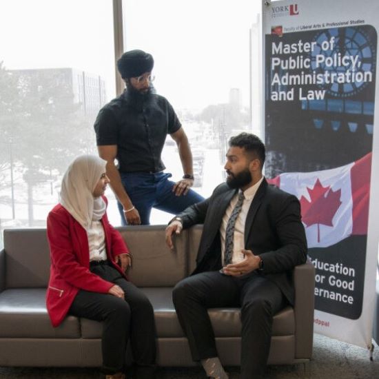 Two professionals sitting on a couch, and one standing behind as they have a discussion together.