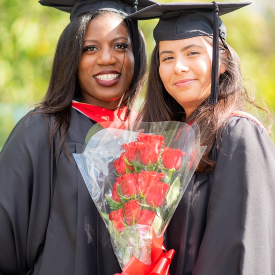 Two smiling graduates in black caps and gowns, one holding a bouquet of red roses, stand together against a backdrop of green foliage.