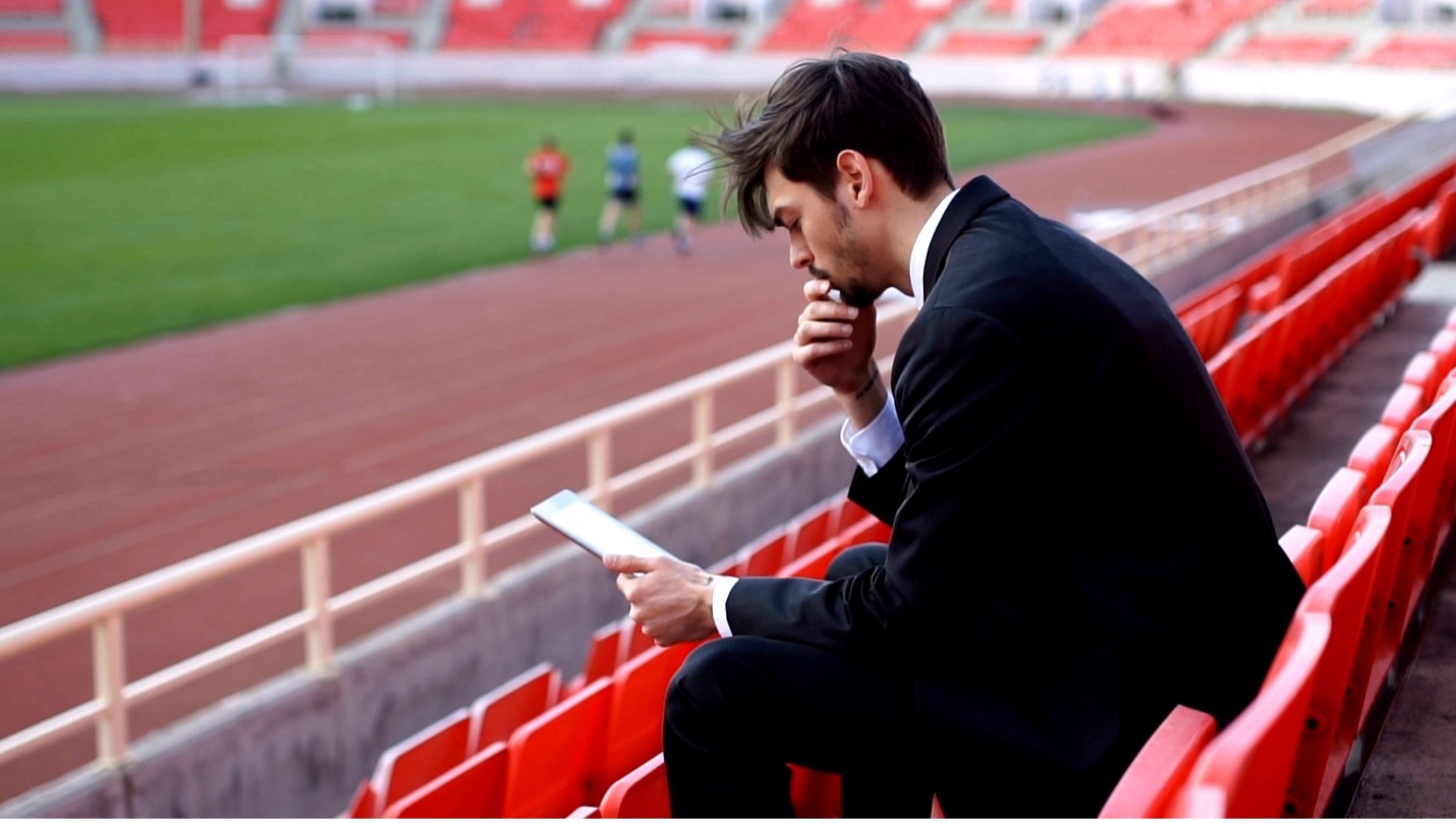 A sport entrepreneur sitting in the stands overlooking sports field looking down at a tablet