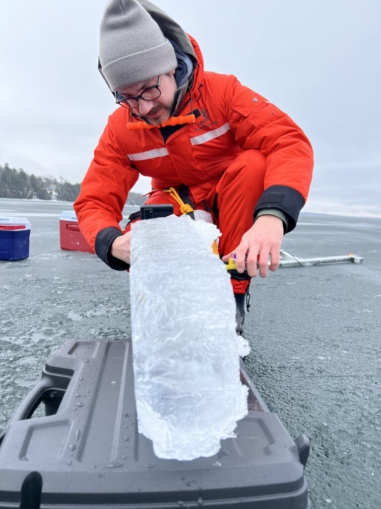 York Postdoctoral Fellow Joshua Culpepper in a red survival suit uses a measuring tape to measure a chunk of ice on Lake Simcoe. Photo by former York Postdoc Kirill Shchapov