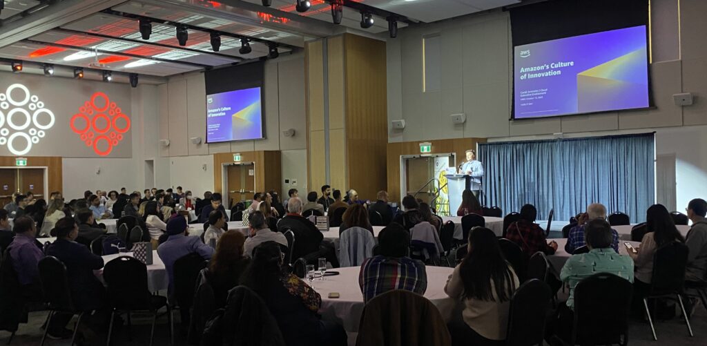 conference with people sitting at round tables and a presenter speaking on stage 