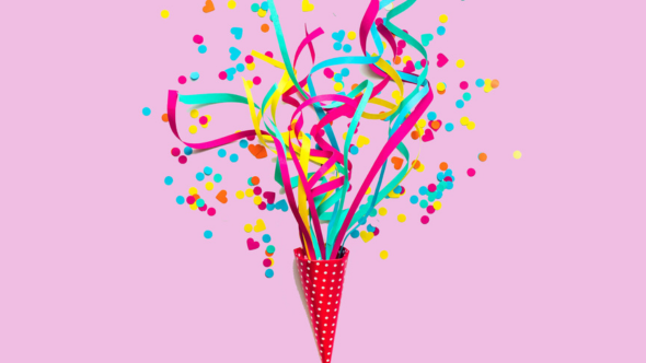 A red polka dot cone with confetti blasting out of it on a pink background