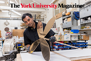 Andil Gosine, a Brown man with dark hair, wearsblack gloves as he holds a metallic flower shaped piece of art in an art studio.