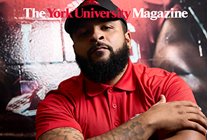 A Black man with a short beard facesthe camera with his arms crossed. He is wearing a buttoned up red gold shirt and black York Lions cap. 