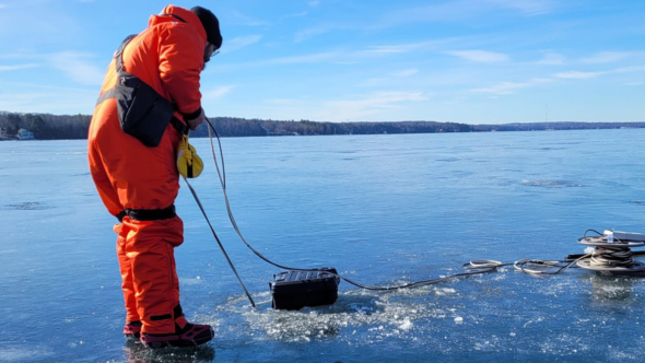 A man in an orage suit standing over an iced out lake holds some rope over a hole in the ice.  