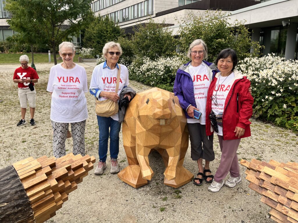 Lynn Taylor, Claudia Hungerson, Gwyn Buck and Grace Chui are pictured in the courtyard of the Rob and Cheryl McEwen Graduate Study & Research Building, flanking a wooden sculpture of a beaver.  Sheila Forshaw stands in the background.